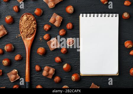 Cioccolato con nocciole, un taccuino con pagine bianche e un cucchiaio di legno con cacao su sfondo scuro, circondato da noci nel guscio e sbucciato. Foto Stock