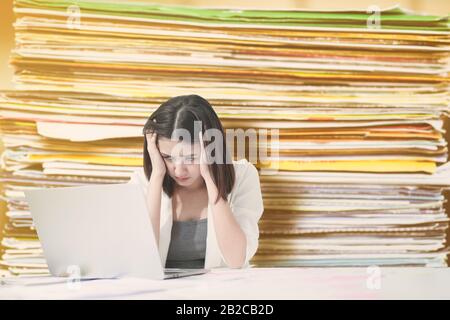 Il bellissimo lavoratore asiatico dell'ufficio sembra stressato e frustrato mentre si lavora digitando e risolvendo il problema sul laptop con un sacco di blurry documento behin Foto Stock