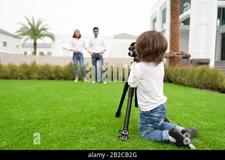 Il retro di un adorabile ragazzo asiatico toddler cercando di scattare una foto del suo genitore con la macchina fotografica sul campo di erba di fronte alla casa, il concetto o Foto Stock
