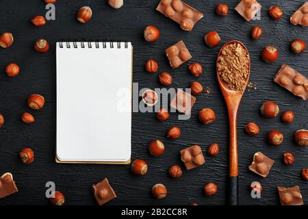 Cioccolato con nocciole, un taccuino con pagine bianche e un cucchiaio di legno con cacao su sfondo scuro, circondato da noci nel guscio e sbucciato. Foto Stock