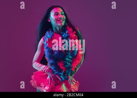 Bellezza. Modello Hawaiian brunette su sfondo studio viola in luce al neon. Belle donne in abiti tradizionali sorridenti, danzanti e divertenti. Vacanze luminose, colori di celebrazione, festival. Foto Stock