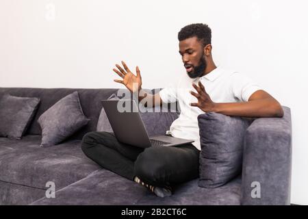 Giovane uomo nero scioccato chattare online su notebook con un amico a casa. Ragazzo informale seduto su un divano beige in un soggiorno leggero, spazio copia Foto Stock