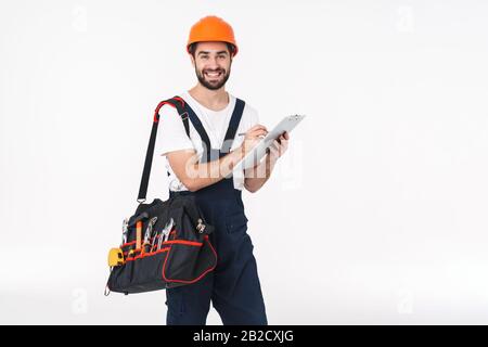 Foto di felice giovane uomo costruttore in casco isolato su parete bianca fondo borsa con attrezzatura strumenti che tiene appunti. Foto Stock