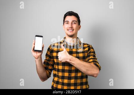 bel giovane uomo o studente con sorriso sul viso tenendo un telefono con schermo bianco verso la fotocamera e mostrando grande pollice in su Foto Stock