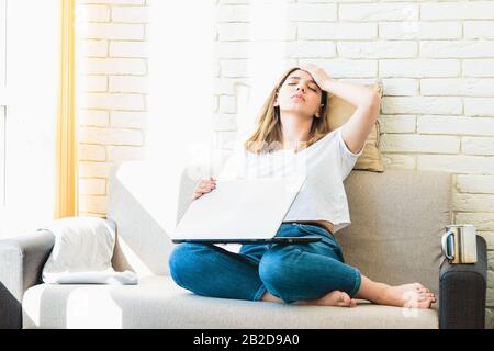 donna stanca sul divano a casa chiudendo il laptop e tenendo la testa Foto Stock