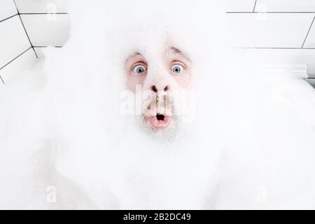 Sorpreso volto maschile in una schiuma lussureggiante, solo gli occhi e la bocca sono visibili, primo piano. L'uomo ama sdraiarsi nella vasca da bagno, con acqua saponata. Foto Stock