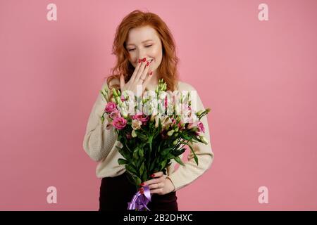 Una ragazza curly dai capelli rossi in un maglione bianco si erge su uno sfondo rosa tenendo un bouquet in mano e sorridendo, nascondendosi dietro il palmo Foto Stock