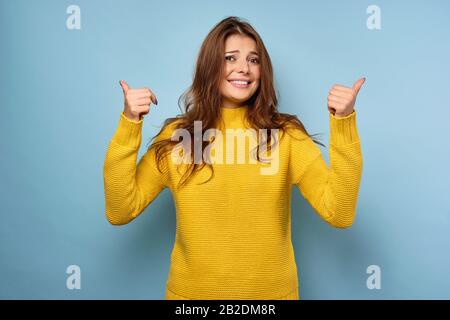 Una bella bruna in un maglione giallo si erge su uno sfondo blu, sorridendo imbarazzato, alzando i pollici in cima Foto Stock