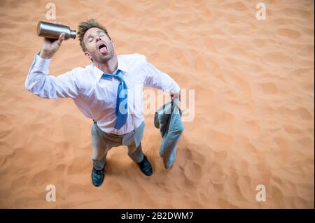 Uomo d'affari assetato in piedi su deserto di sabbia rossa versando una bottiglia d'acqua vuota sopra la sua lingua Foto Stock