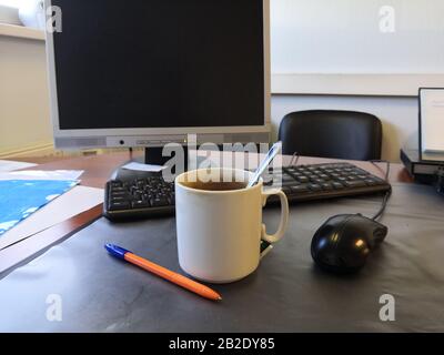 Una tazza di tè sporca su una scrivania da ufficio sullo sfondo di un monitor, una tastiera nera e un mouse nero per computer. Nelle vicinanze si trova una penna a sfera arancione Foto Stock