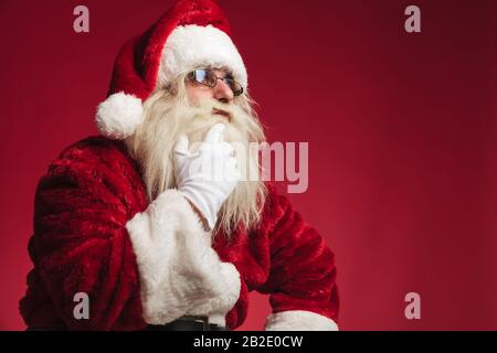 babbo natale che tocca la barba e il mento e si affaccia sullo sfondo rosso Foto Stock