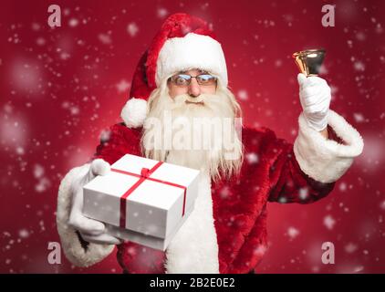 felice babbo natale che suona la sua campana e offre un regalo a tutti mentre nevica su sfondo rosso Foto Stock