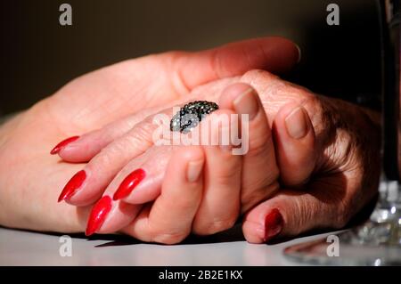 Figlia adulta che tiene la madre stropicciata vecchia mano indossando un anello e smalto rosso del chiodo. Scena parzialmente illuminata con un effetto soft drammatico. Mani sulla tabella Foto Stock