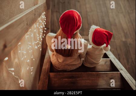 Madre bionda in abiti leggeri e suo figlio in un maglione beige, in cappelli rossi di Natale, sedersi con le spalle su una scala di legno decorata con giallo Foto Stock