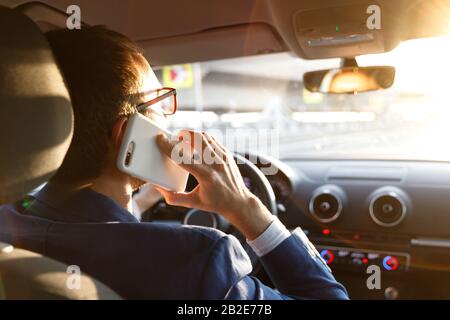 Uomo in blazer utilizzare e parlare sullo smartphone durante la guida di un'auto al tramonto, guardando con attenzione la strada, vista posteriore. Foto Stock