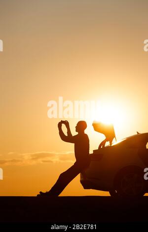 Silhouette di uomo conducente rilassante dopo un giro, seduto sul tronco della sua auto e scatta una foto al tramonto sullo smartphone, vista laterale. Vocazione estiva. Co Foto Stock