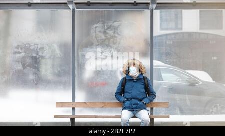 Malato con un cappuccio seduto da solo su panchina di trasporto pubblico, indossando maschera facciale protettiva contro le malattie infettive trasmissibili e come protettori Foto Stock