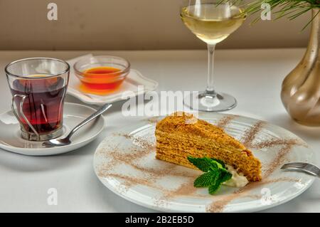 Fetta di torta di miele stratificato. Una rosetta con miele, un bicchiere di vino bianco fresco e una tazza di tè aromatico spesso. Messa a fuoco selettiva. Foto Stock