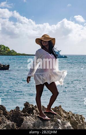 Donna nera da 25 a 30 anni, in piedi su rocce, vestita di modellazione bianca, fresco estate e abiti tropicali, con paesaggio spiaggia e sole s Foto Stock