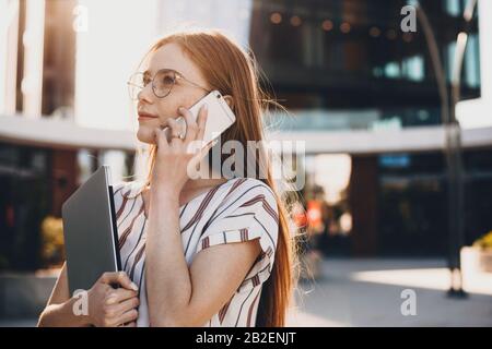 Affascinante donna d'affari con capelli rossi e occhiali che parla al telefono durante una passeggiata fuori dall'ufficio con il suo computer Foto Stock