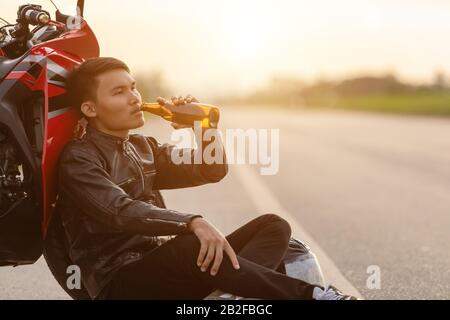 Motociclista seduto sulla strada accanto alla sua moto e bere un alcol o birra. Di sicuro ride concept Foto Stock