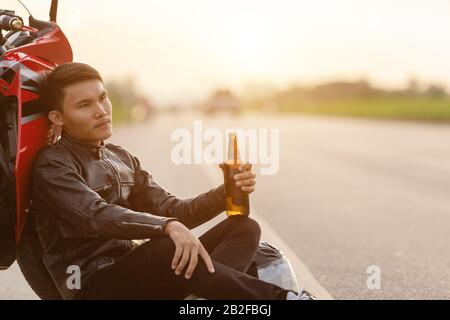 Motociclista seduto sulla strada accanto alla sua moto e bere un alcol o birra. Di sicuro ride concept Foto Stock