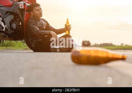 Motociclista seduto sulla strada accanto alla sua moto e bere un alcol o birra. Di sicuro ride concept Foto Stock