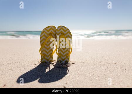 Coppia di infradito a motivo a zig-zag giallo e bianco incastonati nella sabbia su una spiaggia con mare calmo e cielo blu chiaro in estate Foto Stock