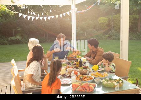 Vista frontale di una famiglia caucasica multi-generazione seduta fuori ad un tavolo da pranzo fissato per un pasto, ascoltando il nonno parlare e sorridere. Foto Stock