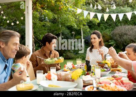 Vista frontale di una famiglia caucasica multi-generazione seduta fuori ad un tavolo da pranzo fissato per un pasto, mangiare, parlare e servire gli altri cibo. Famil Foto Stock