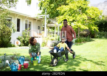 Vista frontale di una felice coppia afroamericana e loro figlio nel giardino, l'uomo spingendo suo figlio in una carriola mentre la madre guarda. Famiglia e Foto Stock