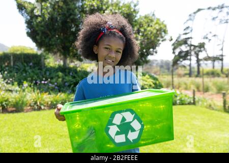 Ritratto di una giovane ragazza afroamericana in piedi in un campo che tiene una cassa verde di riciclaggio platico e sorridente alla macchina fotografica Foto Stock