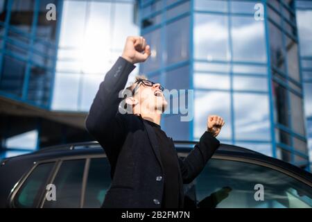 Un giovane uomo di successo, uomini d'affari executive braccia alzate per celebrare il coraggio gridando di fronte a un alto blocco di uffici in una città moderna Foto Stock
