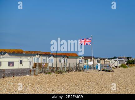 Statico caravan bianco dietro una recinzione con spiaggia di ghiaia e bandiera britannica in pole al West Sands Caravan Park, Bunn Leisure, Selsey, West Sussex, Inghilterra. Foto Stock