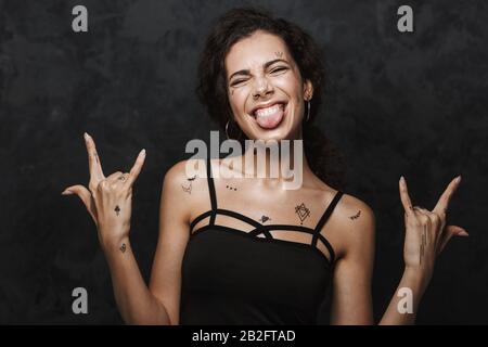 Immagine di giovane donna eccitata con tatuaggio che si steccano la lingua e corna gesturanti con dita isolate su sfondo nero Foto Stock