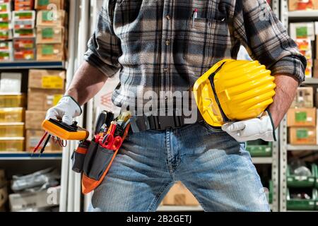 L'elettricista tiene a portata di mano il tester per multimetro, l'elmetto con occhiali di protezione. Industria edile, impianto elettrico. Foto Stock