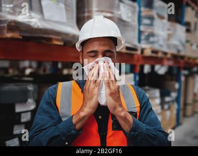 Giovane manager africano che tossisce e si sente male mentre copre la bocca con fazzoletto in magazzino Foto Stock