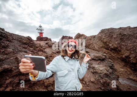 Donna elegante e spensierata che si gode un viaggio su un paesaggio roccioso, facendo foto selfie vicino a un faro Teno a nord-ovest dell'isola di Tenerife Foto Stock