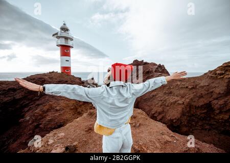 Donna elegante e spensierata vestita di jeans e cappello rosso che si gode un viaggio su una costa rocciosa dell'oceano vicino al faro, viaggiando a nord-ovest dell'isola di Tenerife, Spagna Foto Stock