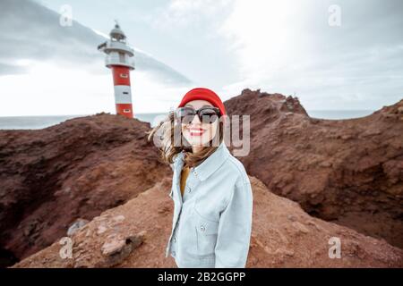 Ritratto di una donna elegante spensierata vestita di jeans e cappello rosso godendo viaggio su una costa rocciosa dell'oceano vicino al faro, viaggiando a nord-ovest dell'isola di Tenerife, Spagna Foto Stock