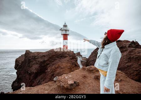 Donna elegante e spensierata vestita di jeans e cappello rosso che si gode un viaggio su una costa rocciosa dell'oceano vicino al faro, viaggiando a nord-ovest dell'isola di Tenerife, Spagna Foto Stock