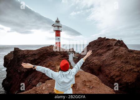 Donna elegante e spensierata vestita di jeans e cappello rosso che si gode un viaggio su una costa rocciosa dell'oceano vicino al faro, viaggiando a nord-ovest dell'isola di Tenerife, Spagna Foto Stock