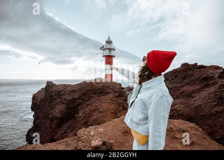 Donna elegante e spensierata vestita di jeans e cappello rosso che si gode un viaggio su una costa rocciosa dell'oceano vicino al faro, viaggiando a nord-ovest dell'isola di Tenerife, Spagna Foto Stock