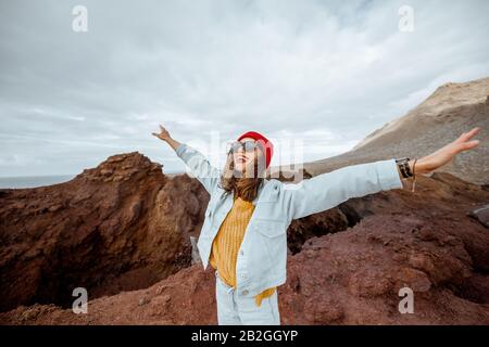 Ritratto di una donna elegante spensierata vestita di jeans e cappello rosso godendo viaggio su una costa rocciosa dell'oceano vicino al faro, viaggiando a nord-ovest dell'isola di Tenerife, Spagna Foto Stock