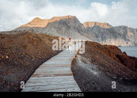 Donna che cammina sul pittoresco sentiero in legno attraverso la terra rocciosa con le montagne sullo sfondo. Viaggiando sul capo nord-ovest dell'isola di Tenerife, Spagna Foto Stock