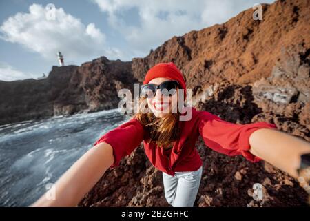 Giovane donna elegante che fa ritratto selfie mentre viaggia sulla costa rocciosa dell'oceano vicino al faro sull'isola di Tenerife, Spagna Foto Stock