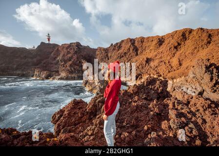 Donna che gode di una splendida vista sulla costa rocciosa dell'oceano e un faro sullo sfondo, viaggiando sul capo Teno a nord-ovest dell'isola di Tenerife, Spagna Foto Stock