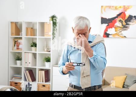 Uomo anziano stanco in camicia blu che tiene gli occhiali e che sfrega il ponticello del naso mentre sente l'affaticamento dell'occhio a casa Foto Stock