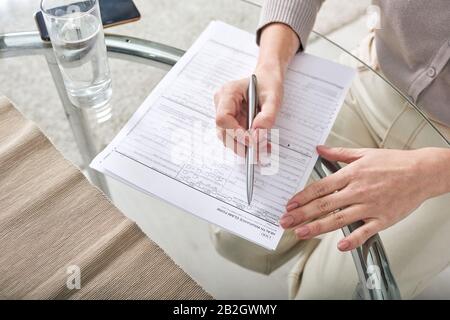 Primo piano di donna irriconoscibile seduto al tavolo vetroso e riempire il reclamo di assicurazione sanitaria a casa Foto Stock