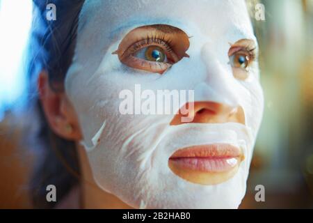 Ritratto di elegante casalinga di 40 anni in pigiama con maschera facciale in foglio bianco sul viso in casa moderna nella soleggiata giornata invernale. Foto Stock